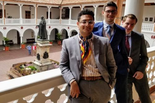 A group of three students in suits stand in the outdoor courtyard of a building